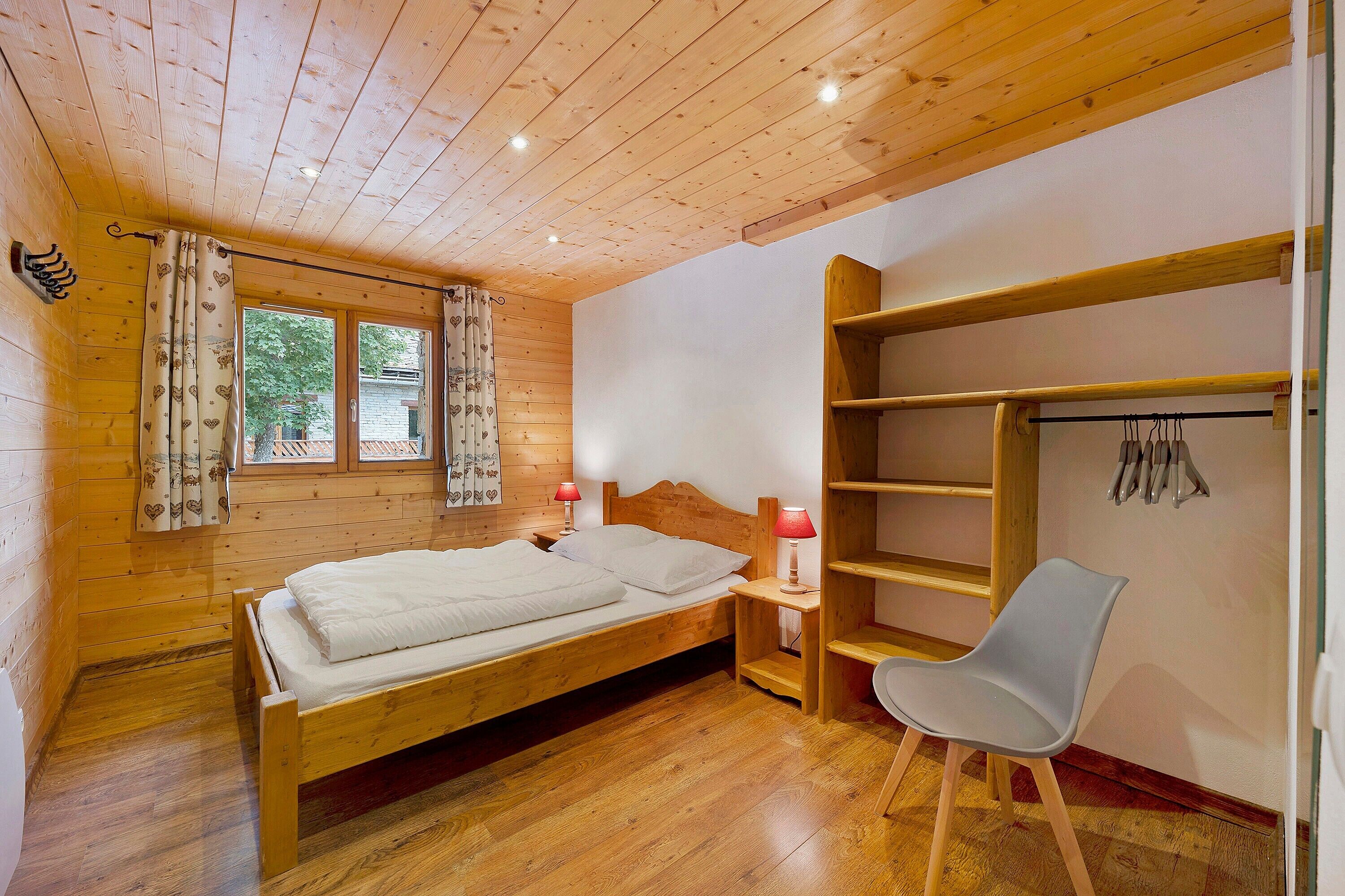 Een kamer met houten vloer, een bed, een stoel, een wandplank en een raam met gordijnen.