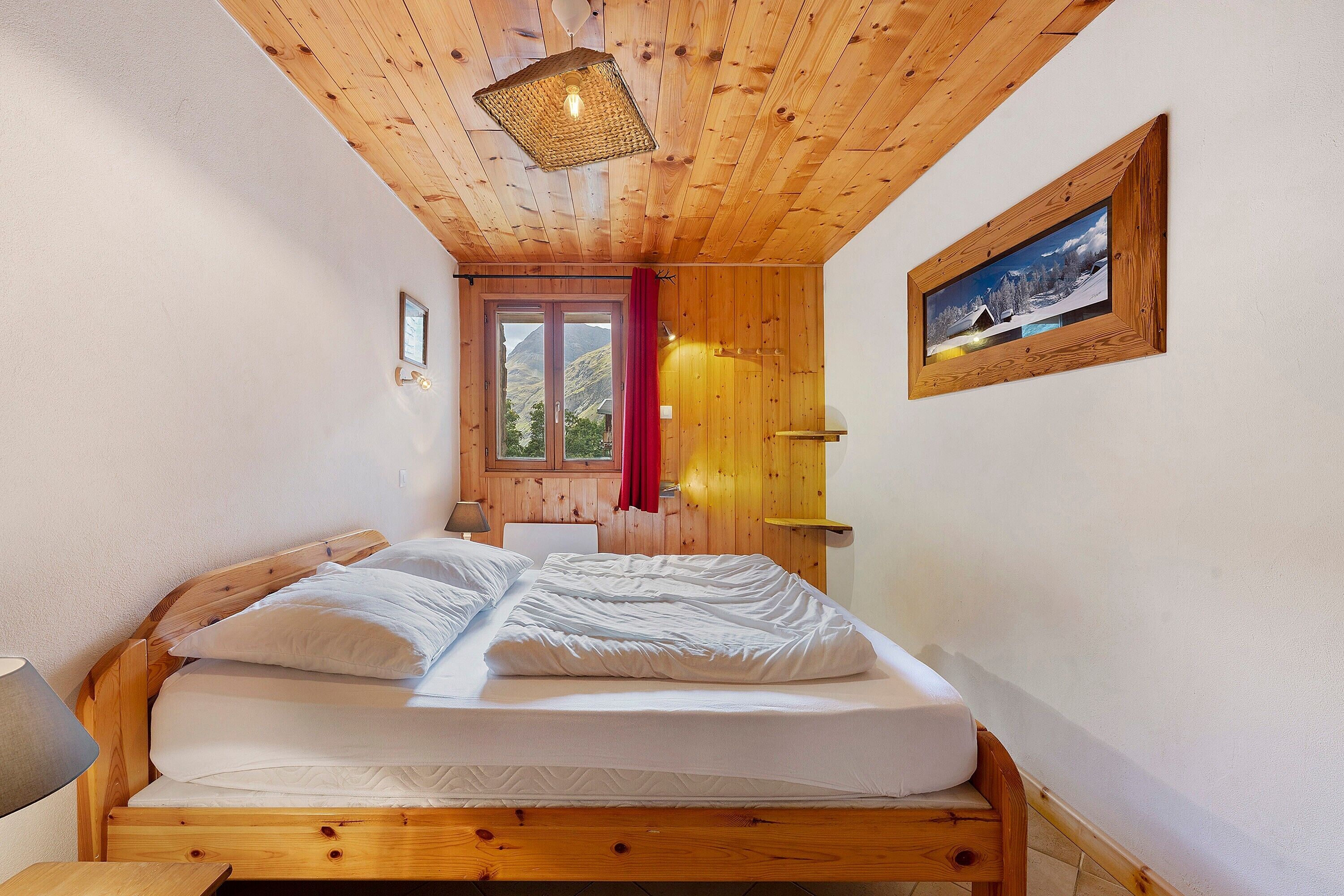 Une chambre avec un plafond en bois, un lit, une fenêtre avec un rideau rouge et un tableau encadré au mur.