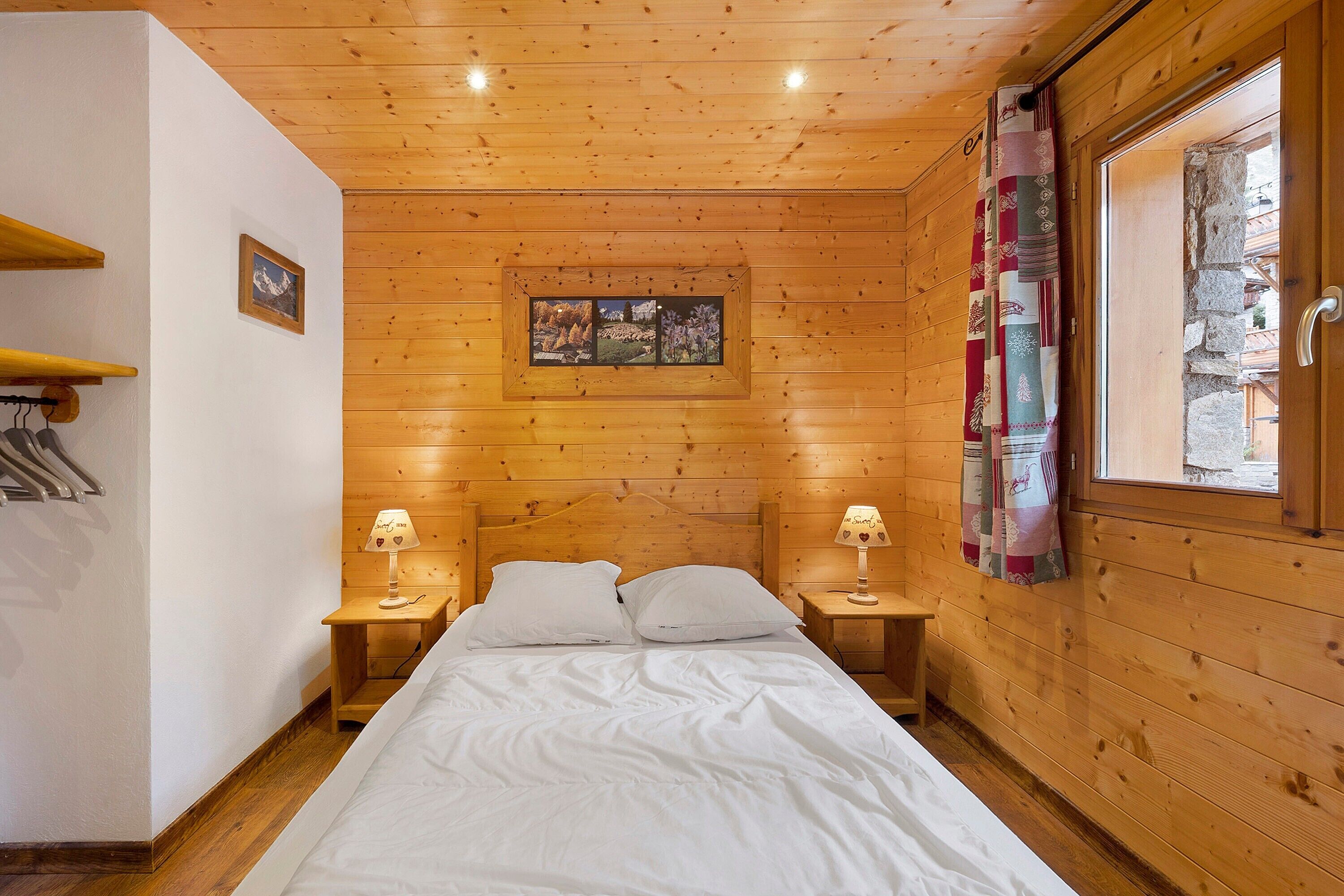 A wooden cabin room with a bed, two bedside tables, a window with curtains, and a picture on the wall.