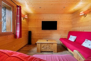 Ein gemütliches Blockhauskabinenzimmer mit Holzwänden, einem Wand-Fernseher, einem kleinen Holztisch, einem roten Sofa und einem Fenster mit Vorhängen.