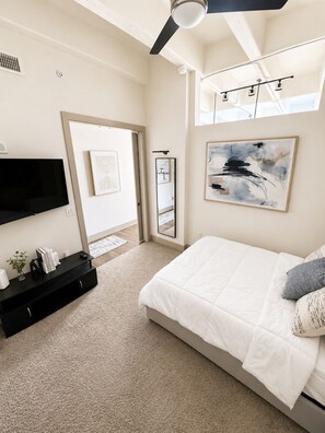 A bedroom with a bed, a flat-screen TV, a ceiling fan, a large abstract painting, and a window. - Luxury High-Rise w/ Skyline View (Atlanta)