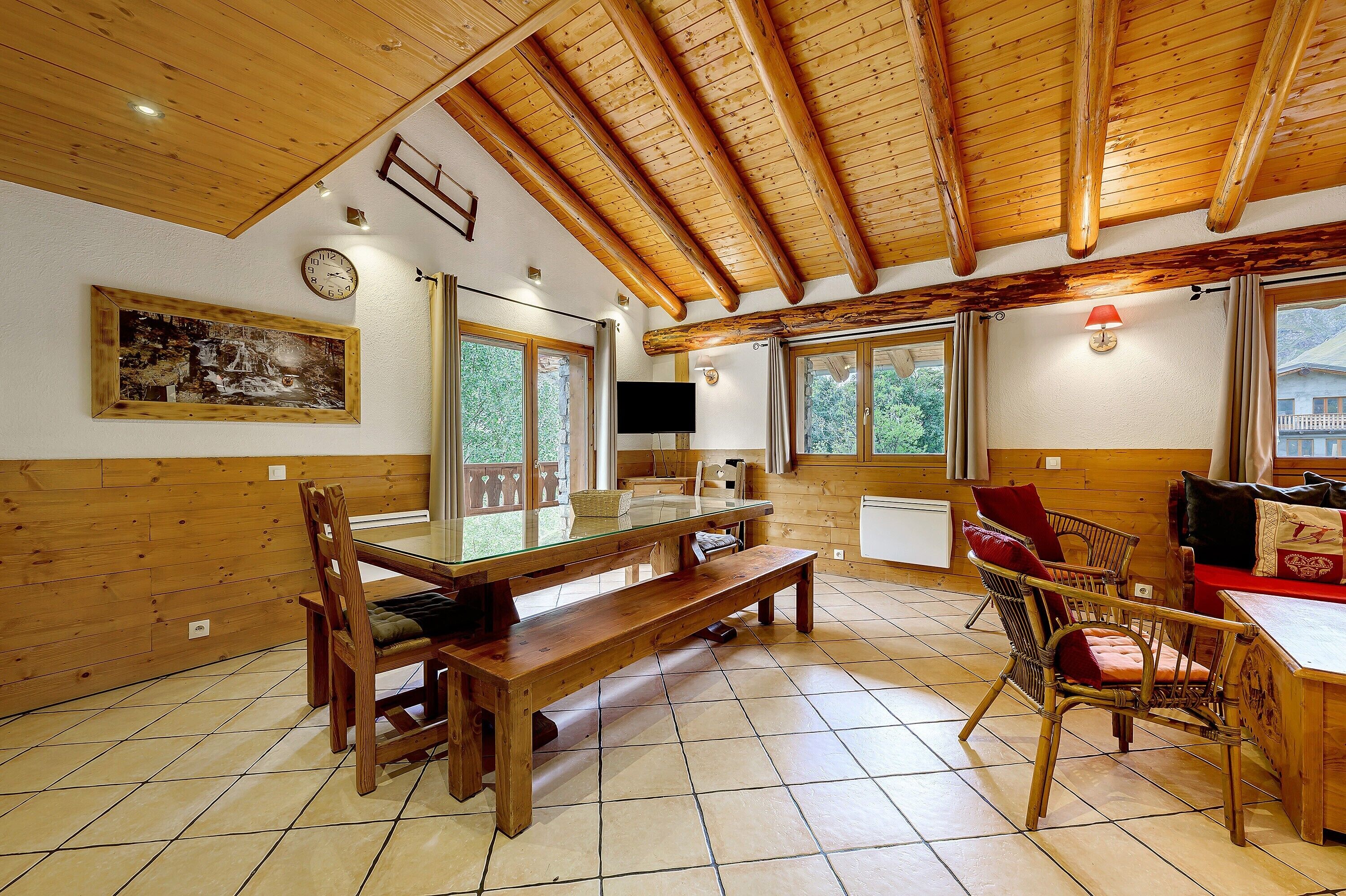 A wooden interior with a dining area, a TV mounted on the wall, and large windows.
