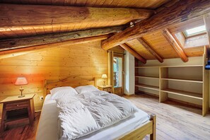 Um quarto de sótão acolhedor, com teto inclinado de madeira, cama com roupa de cama branca, mesa de cabeceira com abajur e estante embutida.