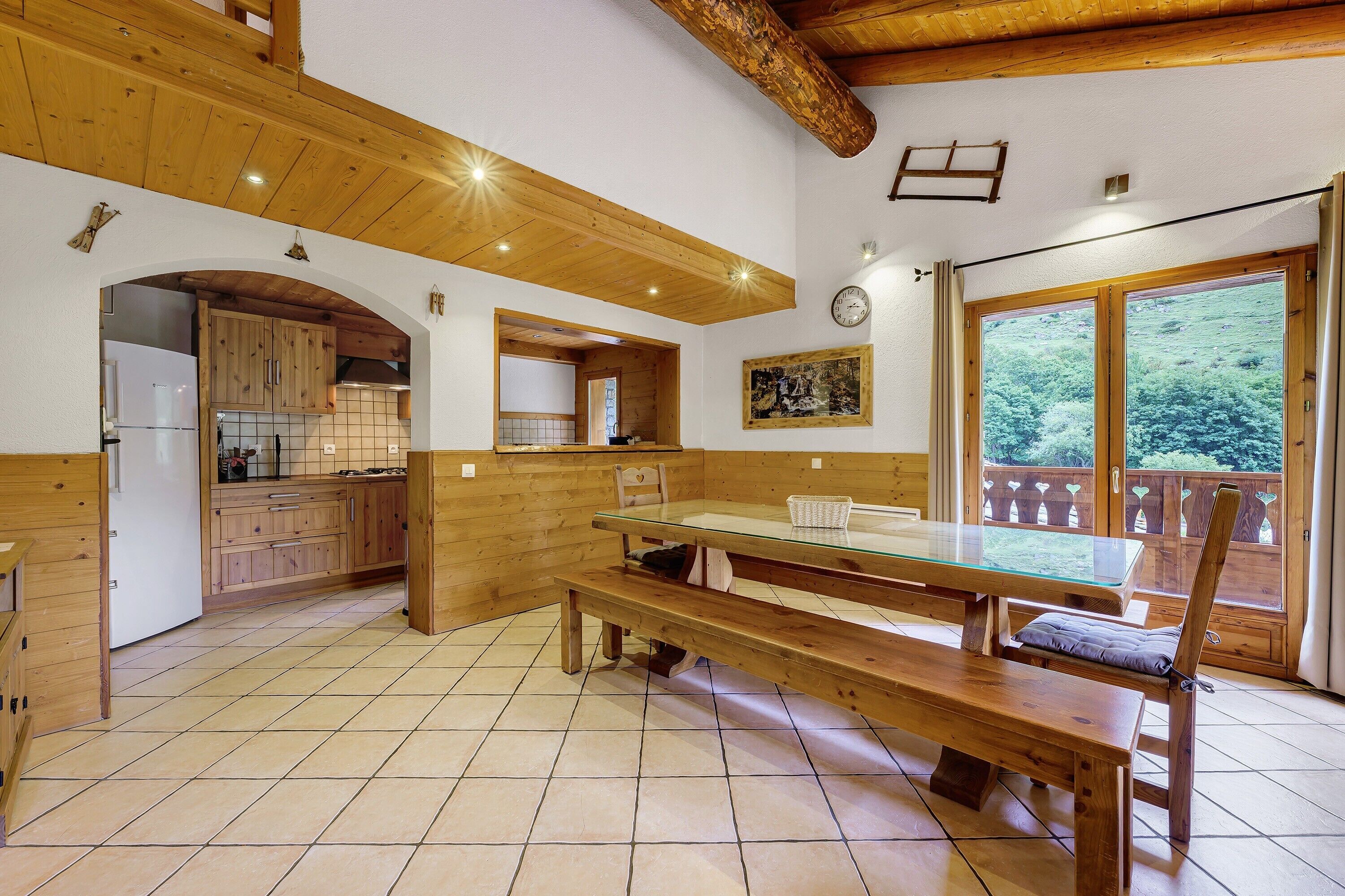 A kitchen with wooden cabinets, a dining table with benches, and a view of greenery through a window.