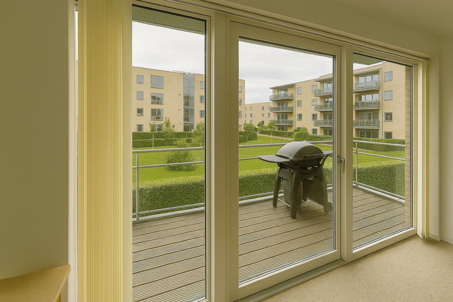 A view from a balcony with a grill and a chair, overlooking a residential area with apartment buildings and a well-maintained lawn.