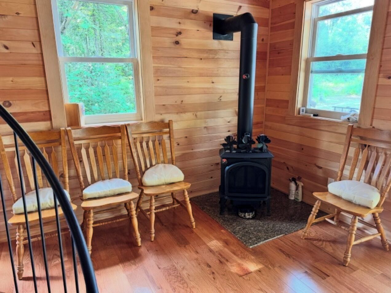 L’intérieur d’un chalet en bois comprend une cheminée, trois chaises en bois avec des coussins et une fenêtre donnant sur un paysage verdoyant.