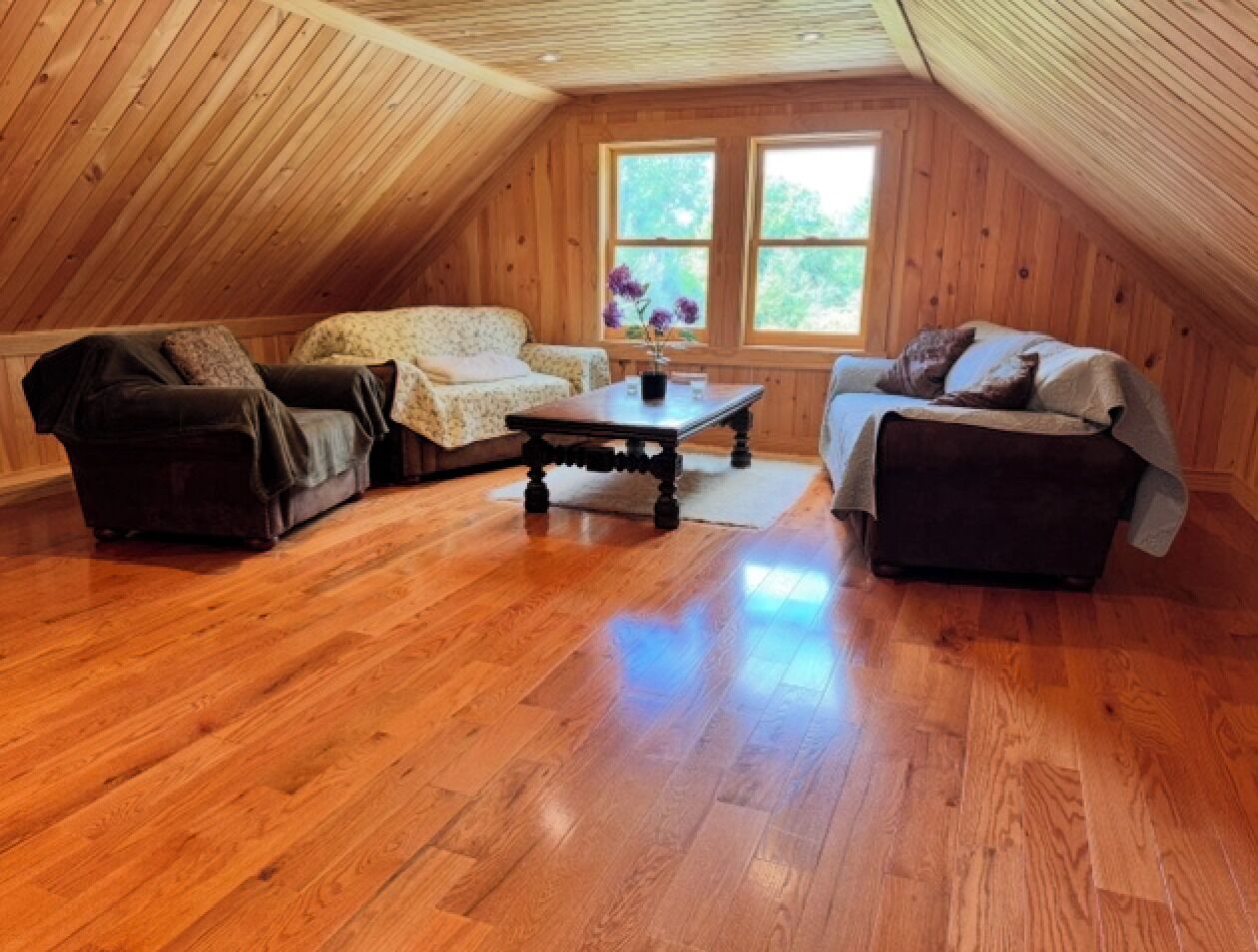 A cozy attic room with wooden flooring, two sofas, a coffee table, and a window with a view of greenery.