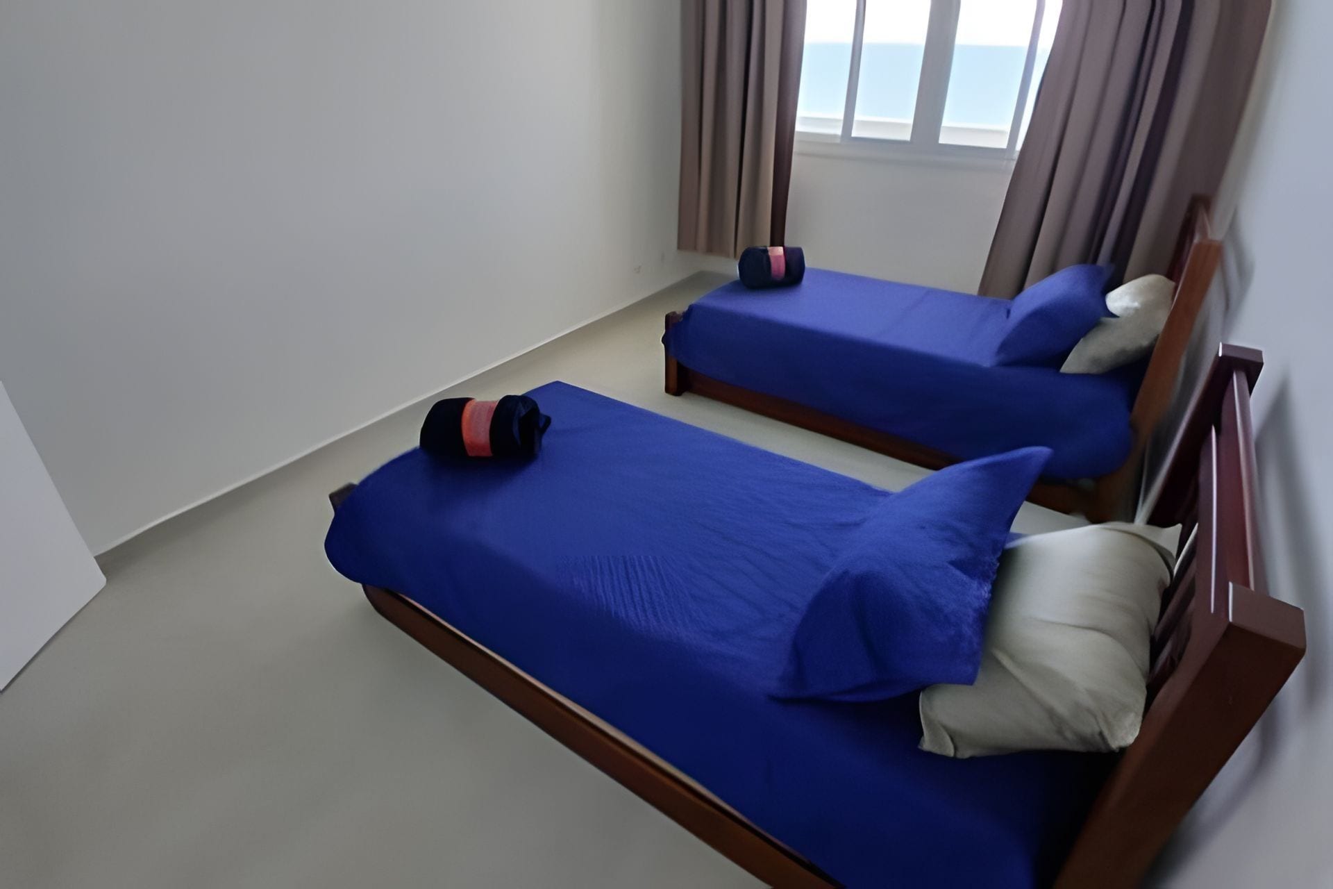 A room with two beds, each with blue bedding and pillows, a wooden headboard, and a window with curtains.