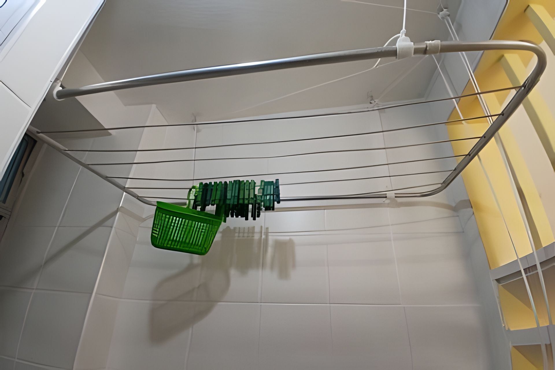 Una cesta de ropa verde colgada de una estructura metálica en una habitación con paredes de azulejos blancos y una pared de acento amarilla.