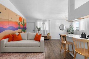 Ein modernes Wohnzimmer mit einer Couch, einem Esstisch und Stühlen. An der Wand ist ein farbenfrohes Wandbild, eine Pflanze und ein Fenster mit Vorhängen.