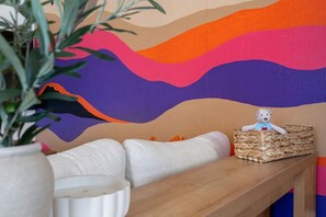 Ein Zimmer mit einem farbenfrohen, abstrakten Wandbild, einer weißen gepolsterten Bank, einem gewebten Korb mit einem Stofftier und einer Topfpflanze.
