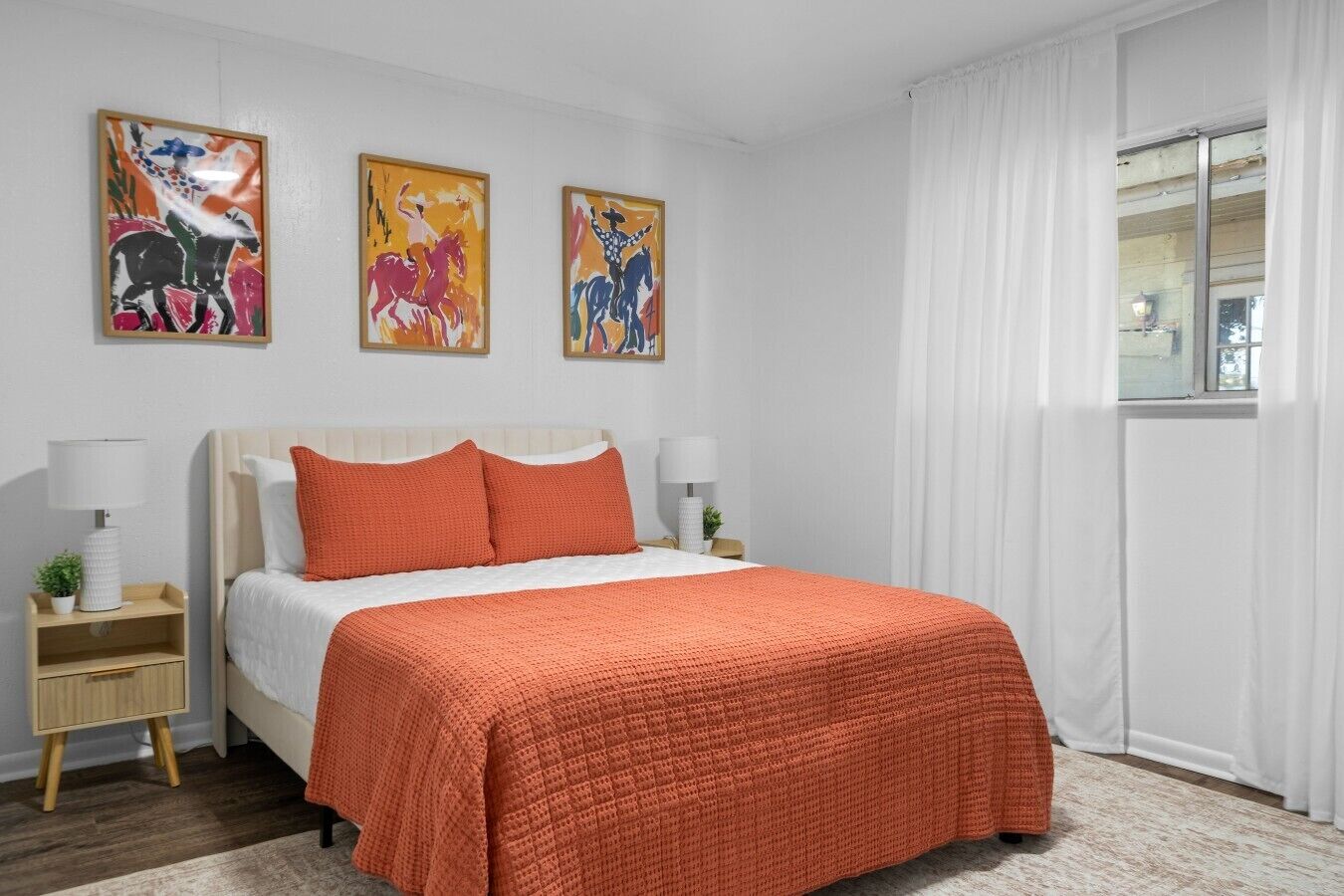 Une chambre à coucher comprenant un lit, deux tables de chevet, une fenêtre avec des rideaux et trois œuvres d’art accrochées au mur.