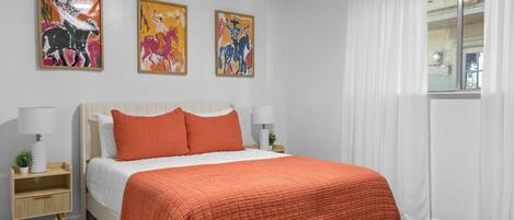 A bedroom with a bed, two bedside tables, a window with curtains, and three wall-mounted artworks.