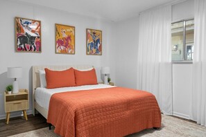 Ein Schlafzimmer mit einem Bett, zwei Nachttischen, einem fensterbelaubten Fenster und drei Wandbildern.