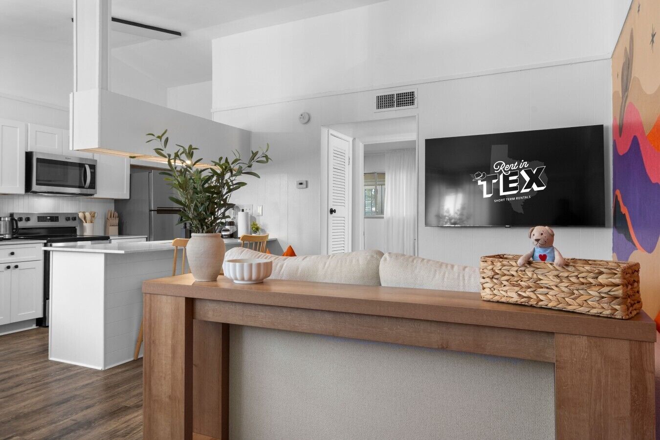 A modern kitchen with white cabinetry, a microwave, and a stove. There is a wooden island with a plant and decorative items. A television is mounted on the wall, and a stuffed toy is on the island.