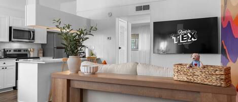 A modern kitchen with white cabinetry, a microwave, and a stove. There is a wooden island with a plant and decorative items. A television is mounted on the wall, and a stuffed toy is on the island.