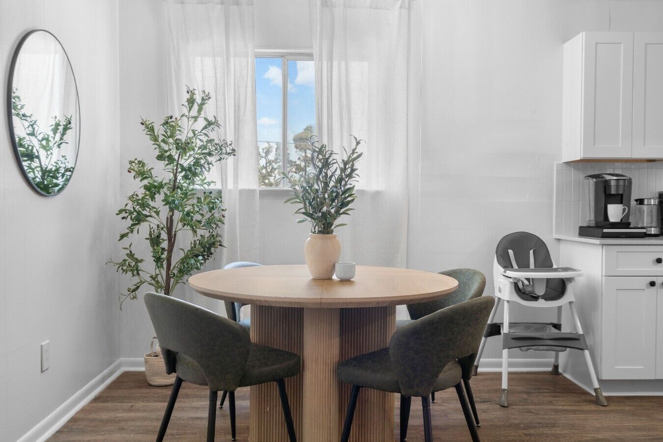 A modern kitchen with white cabinets, a round dining table with chairs, a coffee maker, and a potted plant.