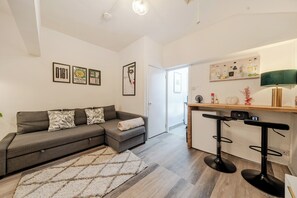 A modern living room with a grey sofa, a small rug, and a kitchen area with a bar stool and a fridge. - Deluxe Apartment5- City Centre Manchester (Manchester)