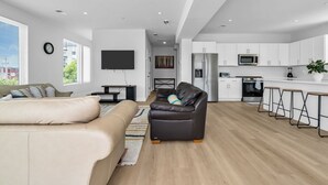 Ein modernes Wohnzimmer mit einer Couch, einem Ledersessel und einem Fernseher. Es gibt eine Küchenzeile mit weißen Schränken und einer Mikrowelle.