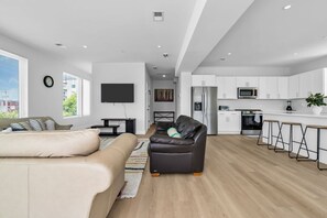Ein modernes Wohnzimmer mit einer Couch, einem Ledersessel und einem Fernseher. Es gibt eine Küchenzeile mit weißen Schränken und einer Mikrowelle.
