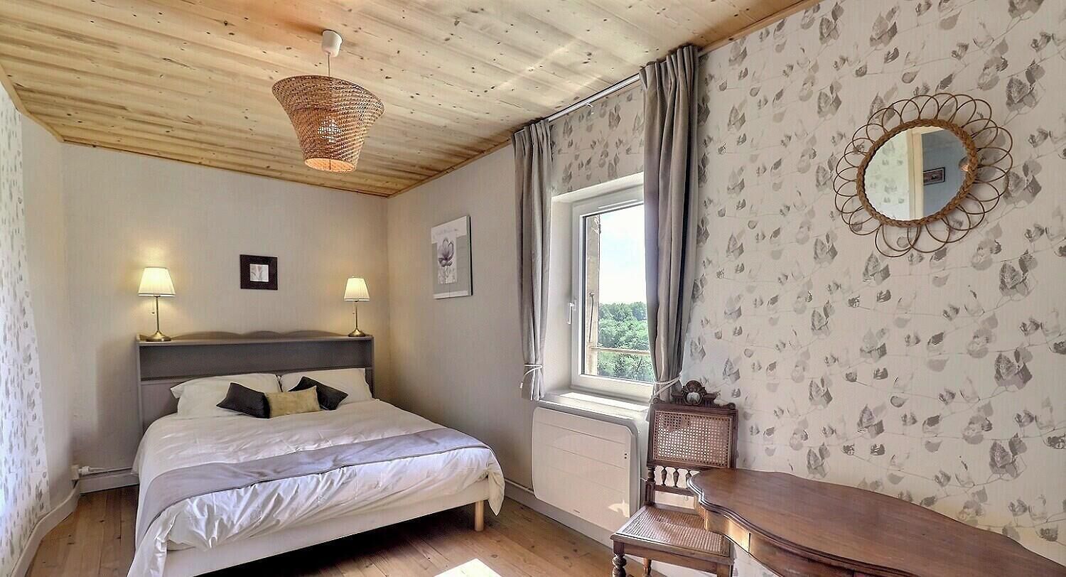 Un dormitorio con cama, lámparas de noche, una ventana con cortinas, una silla de madera y un espejo en la pared.
