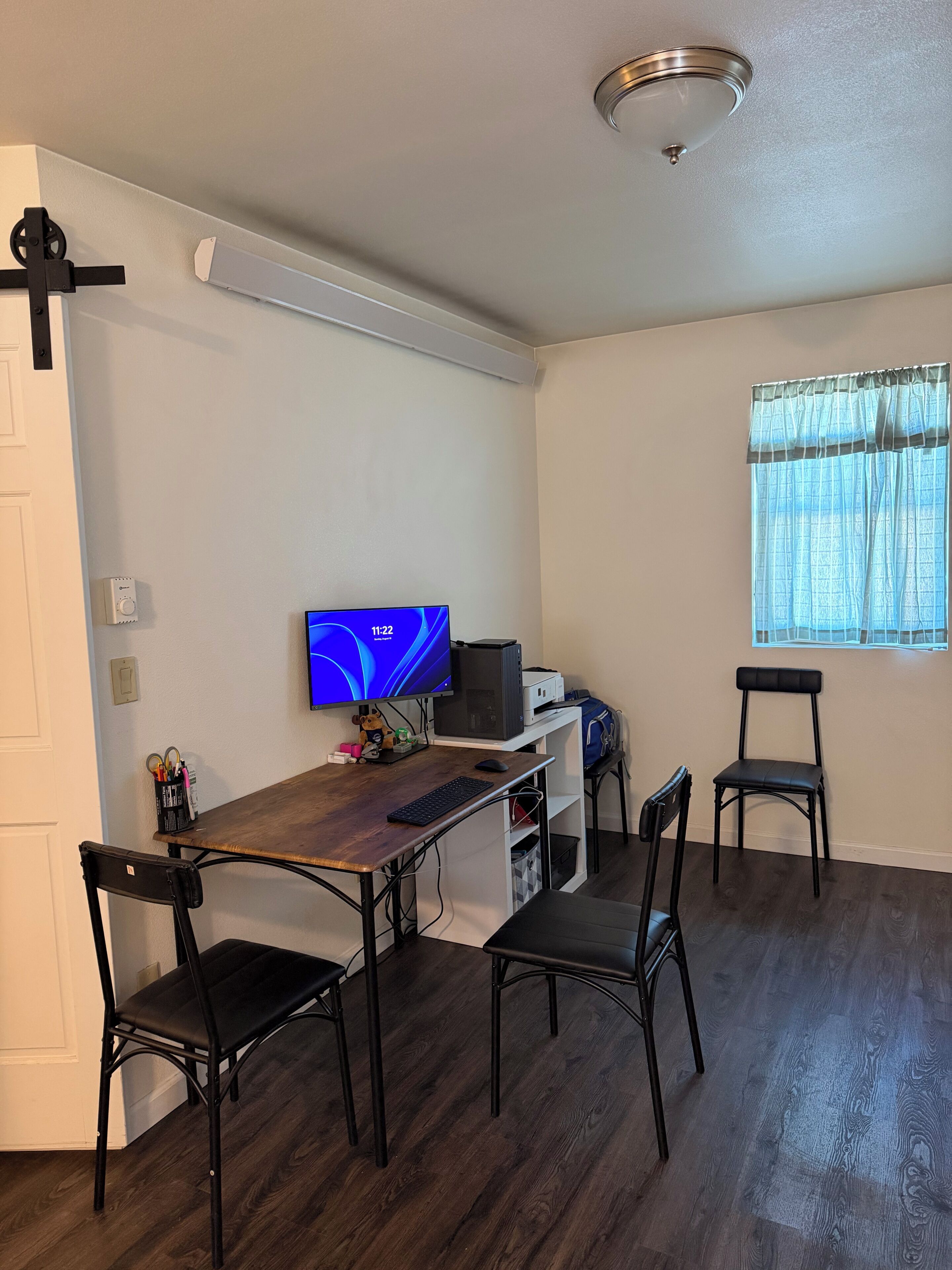 A small, modern room with a wooden desk, two black chairs, a computer, and a window with sheer curtains.