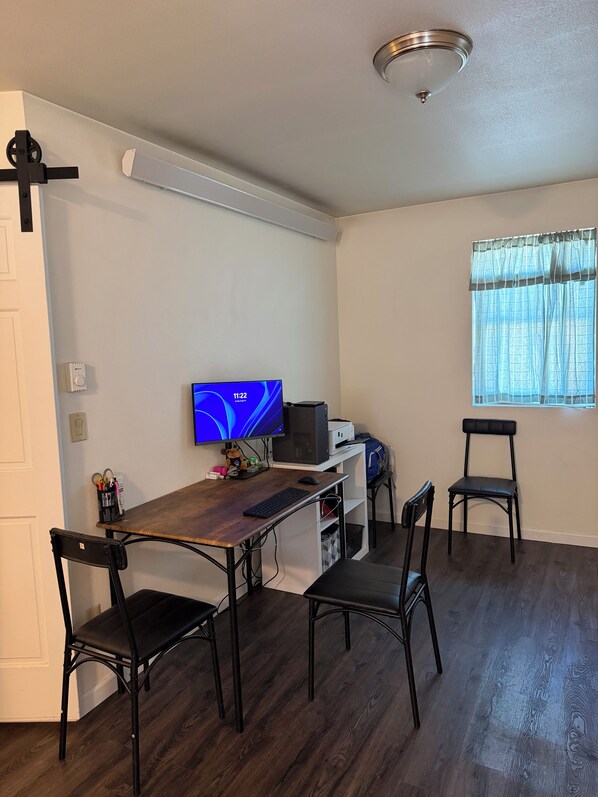 A small, modern room with a wooden desk, two black chairs, a computer, and a window with sheer curtains.