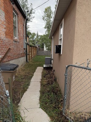 A narrow concrete pathway flanked by a brick building and a stucco building with a chain-link fence.