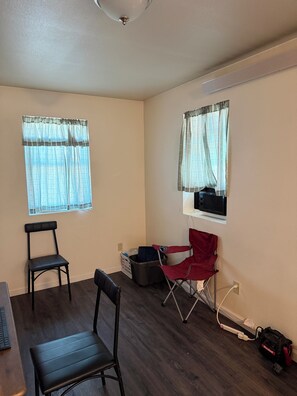 Ein Zimmer mit zwei Fenstern, einem Sessel, einem Schreibtisch und einem roten Klappstuhl.
