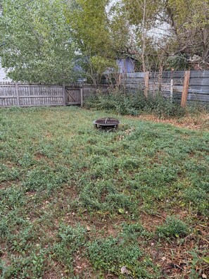 A backyard with a metal fire pit surrounded by grass and weeds.