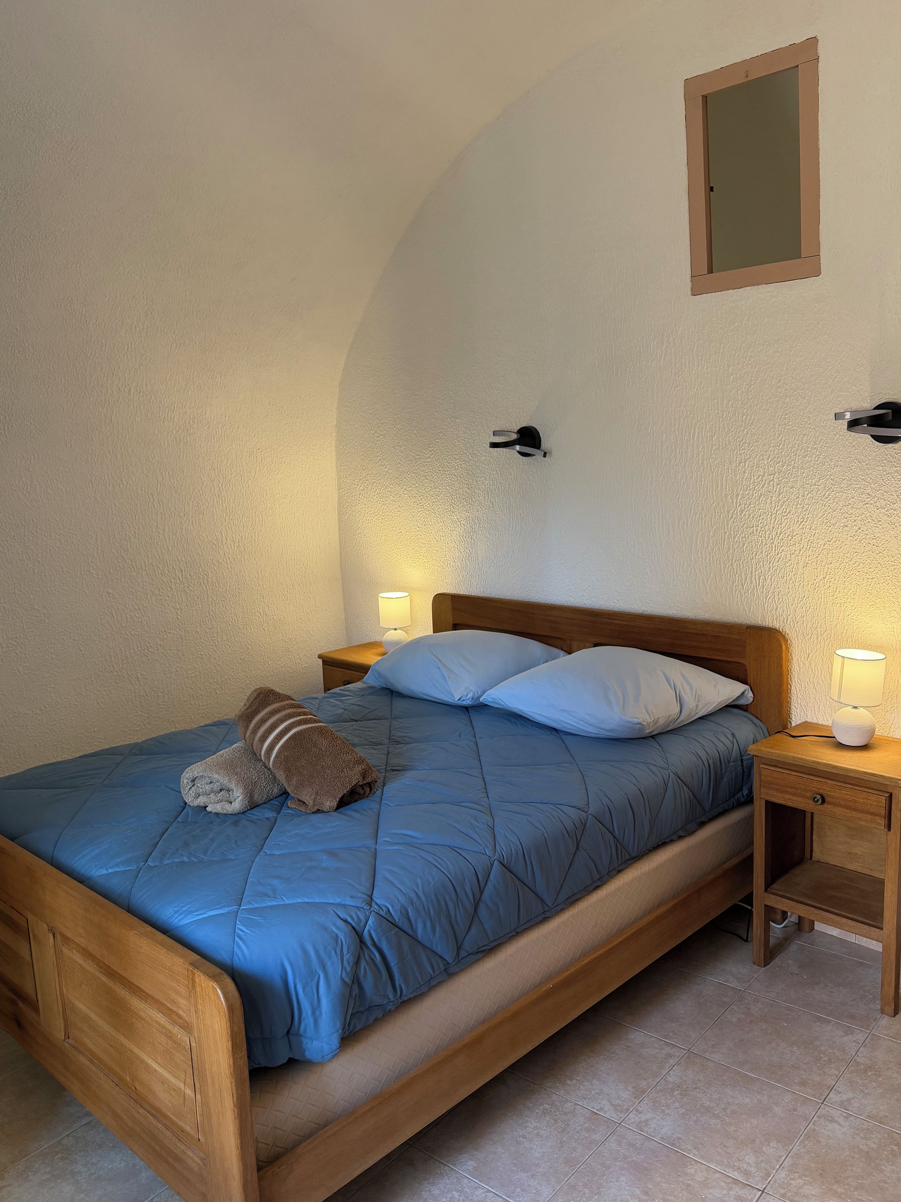 A bedroom with a wooden bed, two bedside lamps, a wall-mounted light, and a small bedside table.