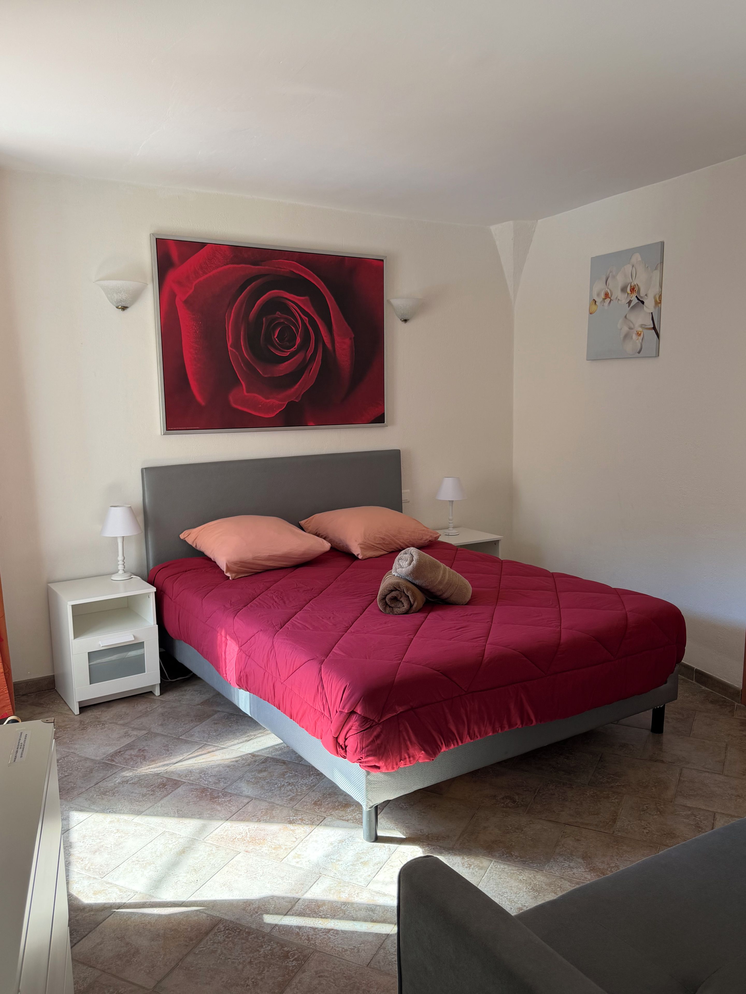 A bedroom with a bed, a nightstand, a wall-mounted lamp, and a painting of a rose.
