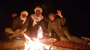 Four individuals gathered around a campfire with a bottle and a plate on the ground. - Authentic Sahara Desert Camp in Mhamid - Peaceful andUnforgettables Stay (Mhamid)