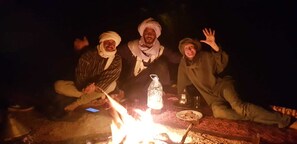 Four individuals gathered around a campfire with a bottle and a plate on the ground. - Authentic Sahara Desert Camp in Mhamid - Peaceful andUnforgettables Stay (Mhamid)