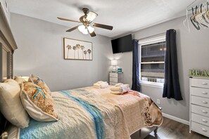 Ein Schlafzimmer mit einem Bett, einem Deckenventilator, einem Fernseher, einem Fenster mit Vorhängen und einer Kommode.