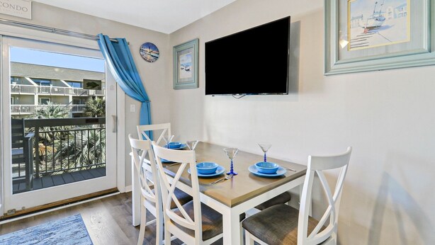 A dining area with a table set for four, a flat-screen TV mounted on the wall, and a balcony with a view of other buildings. - Breakers 220 Oceanfront Condo Direct Beach Access Elevator (Hilton Head Island)