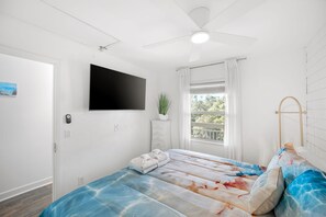 Un dormitorio con cama, ventilador de techo, televisor montado, ventana con cortinas y una planta.