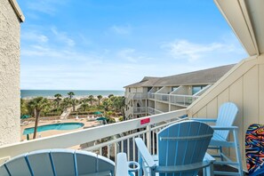 Balcón con vista a la playa y una piscina, con sillas Adirondack azules y un letrero que indica que no se permite asar a la parrilla.