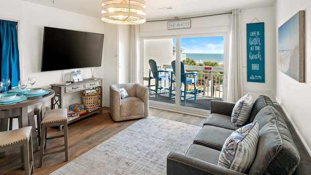 Una sala moderna con televisión de pantalla plana, sofá, comedor con mesa y sillas, y balcón con vista a la playa.