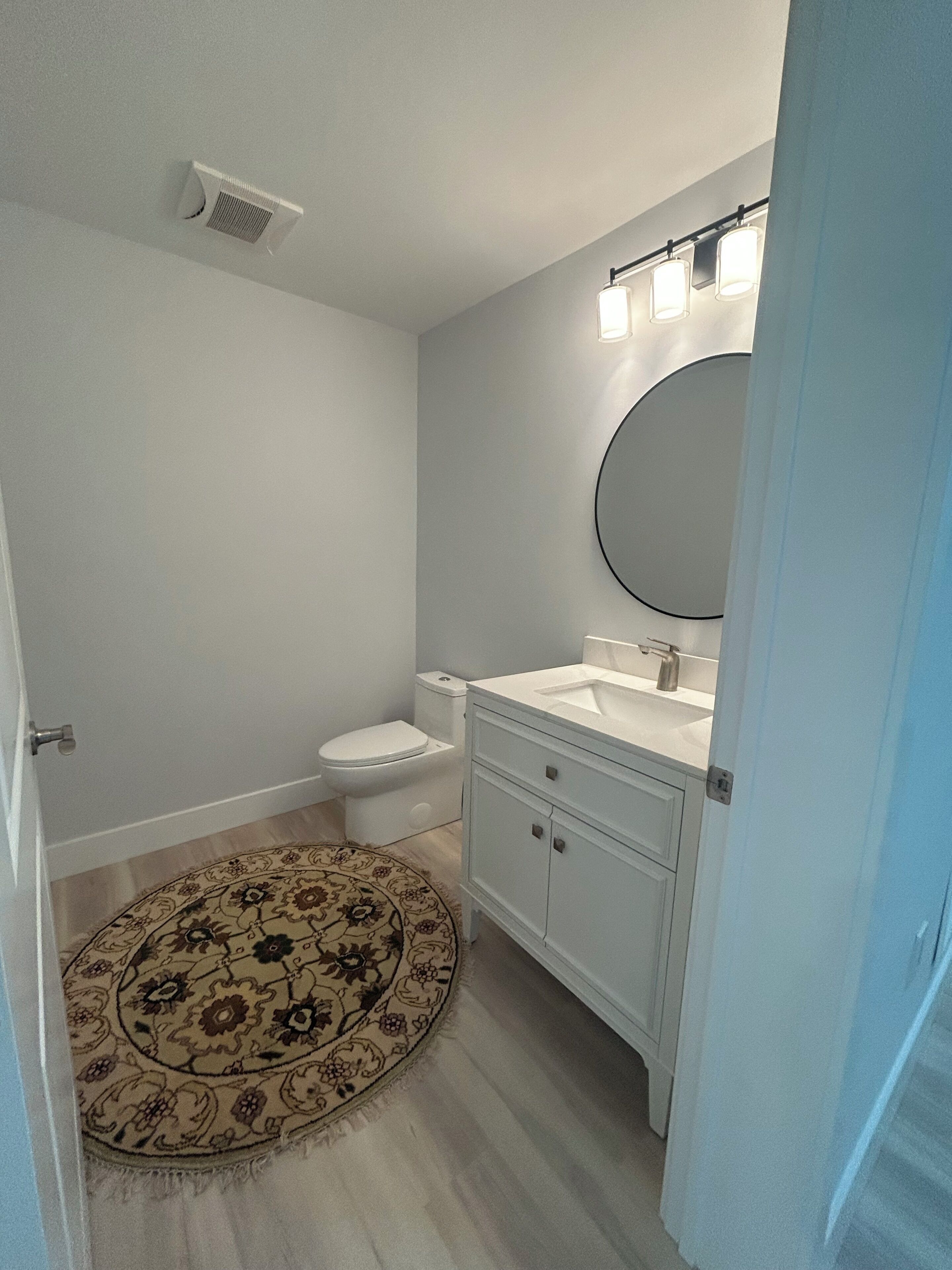 A bathroom with a toilet, sink, mirror, and a patterned rug.