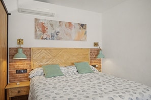 Une chambre avec une tête de lit en briques, un lit recouvert d’une literie à motifs, deux lampes fixées au mur et un grand tableau abstrait.