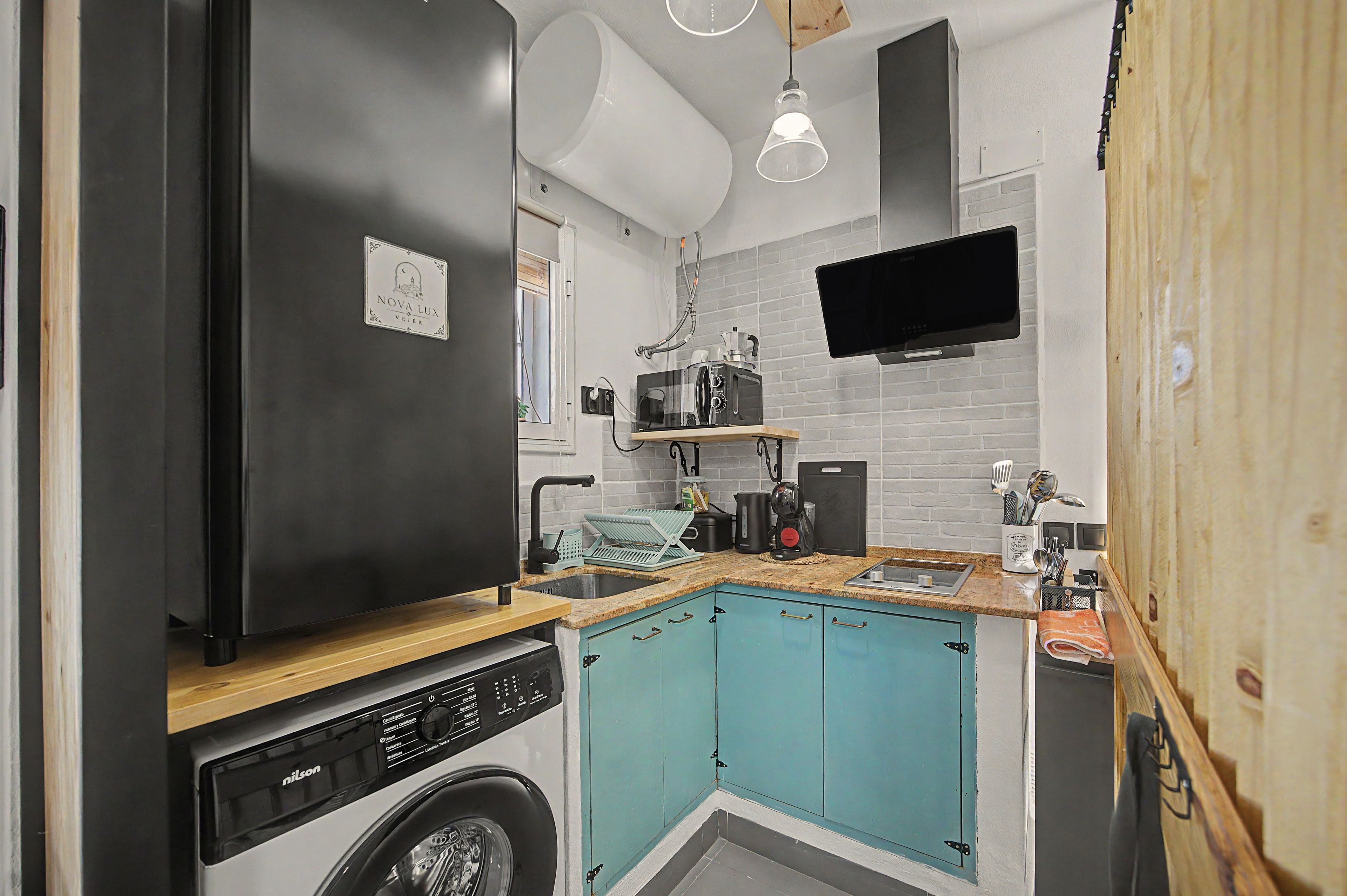 Een compacte keuken met een zwarte koelkast, een wasmachine, blauwgroene kastjes, een magnetron en een televisie aan de muur bevestigd.