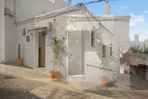Un bâtiment blanc avec une porte, des fenêtres et un petit balcon. On peut voir des plantes en pot et un scooter garé à l’extérieur.