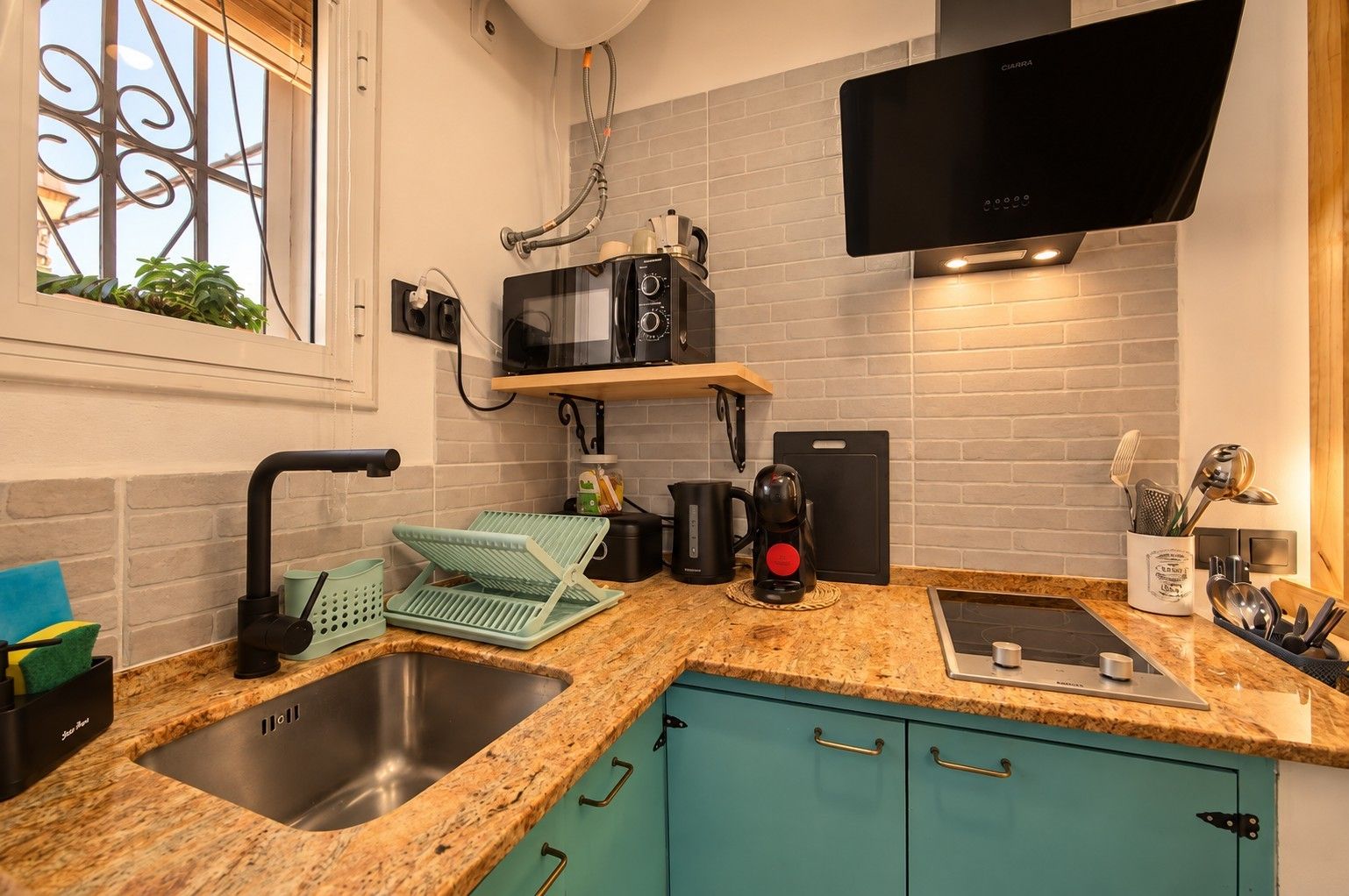Une cuisine moderne dotée d’un comptoir en granit, d’un évier en acier inoxydable, de meubles de couleur turquoise, d’un micro-ondes noir et d’un téléviseur à écran plat fixé au-dessus du comptoir.
