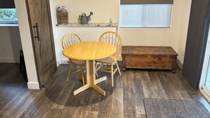 Un comedor con una mesa y sillas de madera, un banco pequeño, una ventana con persianas y una estantería con objetos.