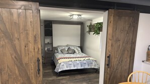 Una habitación con una cama, una puerta de madera, una mesita y una planta colgante.
