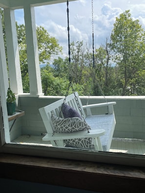 Une véranda avec une chaise suspendue et une petite table, donnant sur un paysage verdoyant et luxuriant.