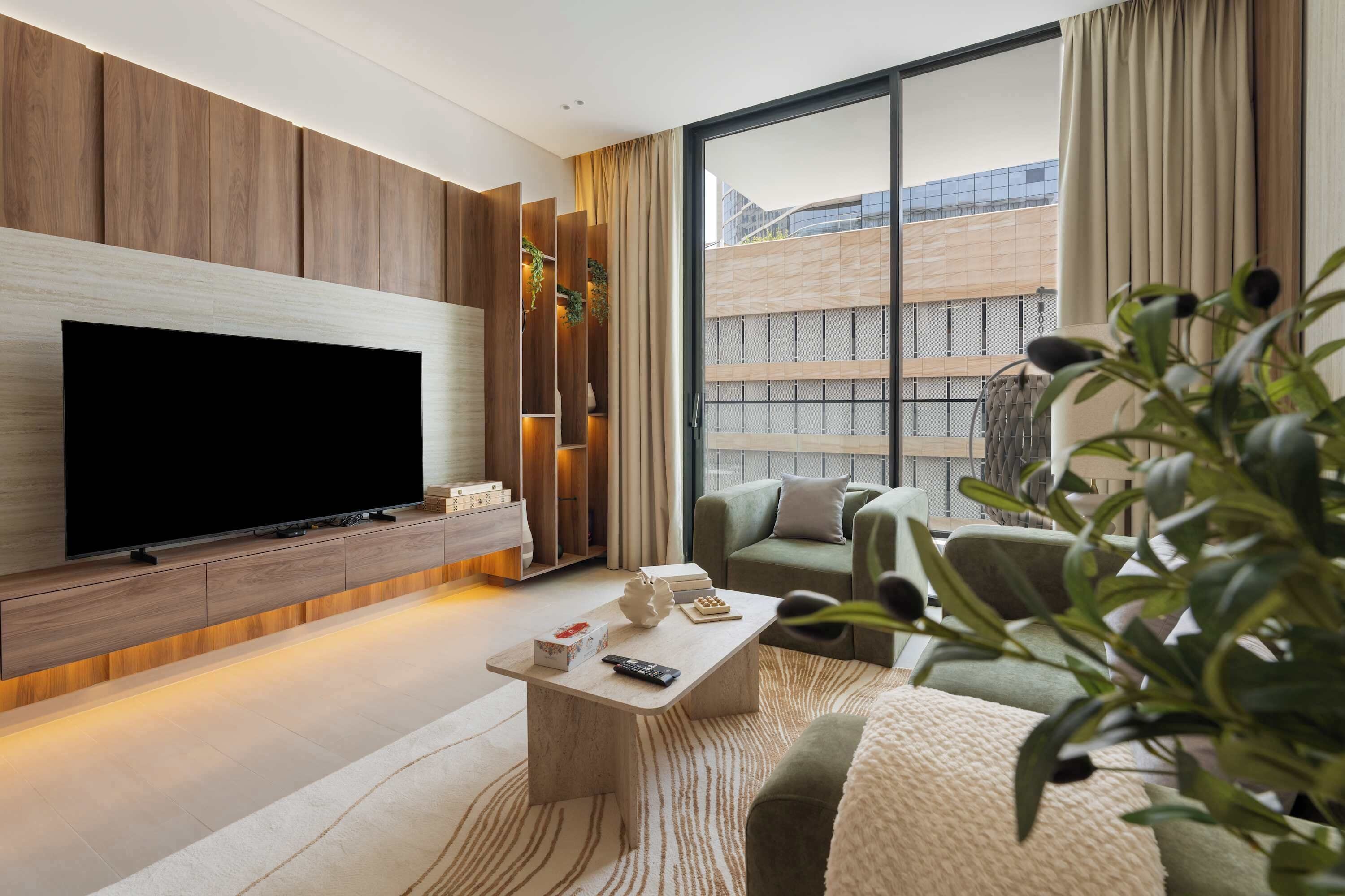 A modern living room with a flat-screen TV mounted on a wooden wall, a sofa, a coffee table, and a large window with curtains.