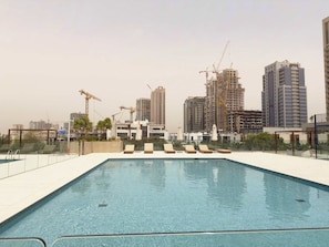 A rooftop swimming pool with lounge chairs and a view of a cityscape with construction cranes. - Stylish 1BR Dubai with Pool, Gym, Padel & Balcony (Jumeirah Village Circle)