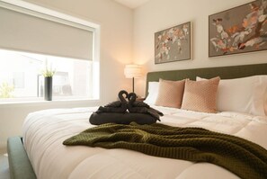 Ein ordentlich gemachtes Bett mit grüner Decke, zwei schwanenförmigen Kissen und einer Lampe daneben.